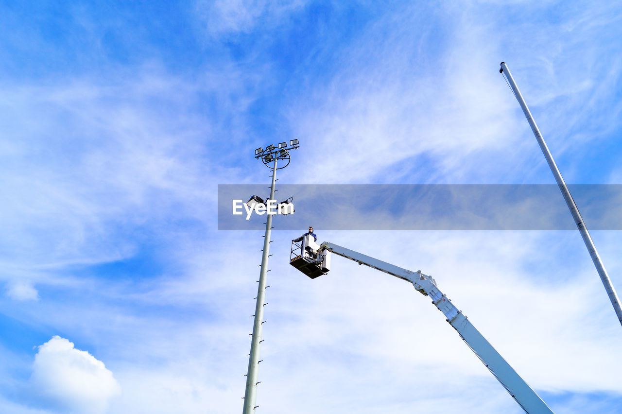 Low angle view of man standing on cherry picker by floodlight against sky
