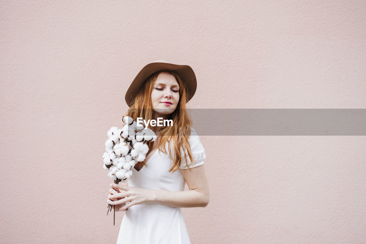 Young woman with eyes closed holding bunch of cotton plant in front of pink wall