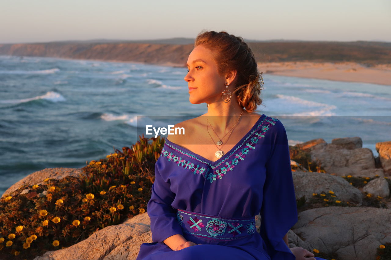 Woman sitting on rock looking at sea shore against sky