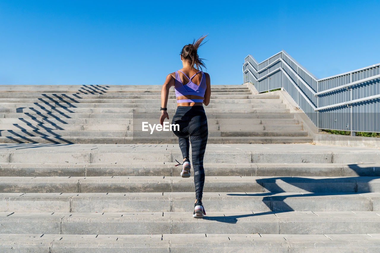 Low angle back view of unrecognizable fit female in activewear running up stone staircase in city during dynamic cardio training