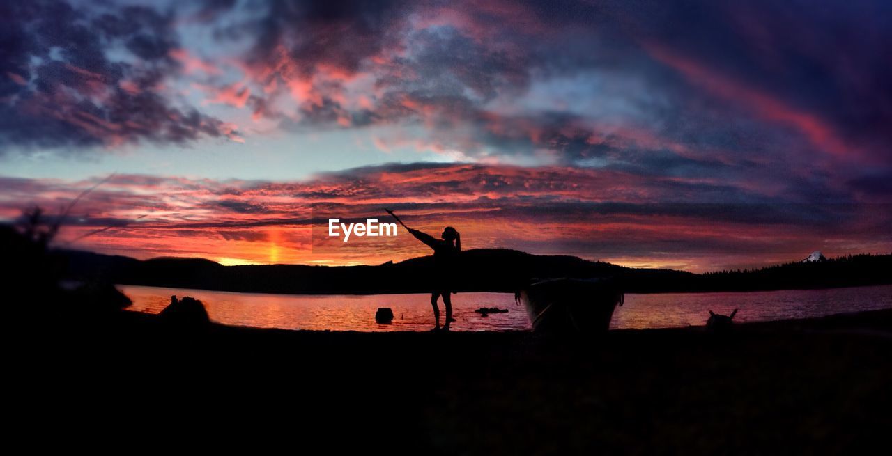 Silhouette woman standing against river and cloudy sky during sunset