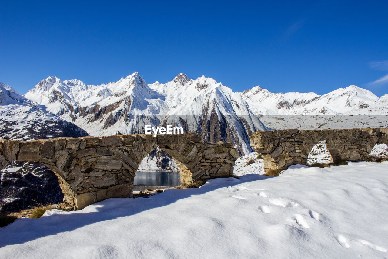 Broken arch bridge by snowcapped mountains against blue sky