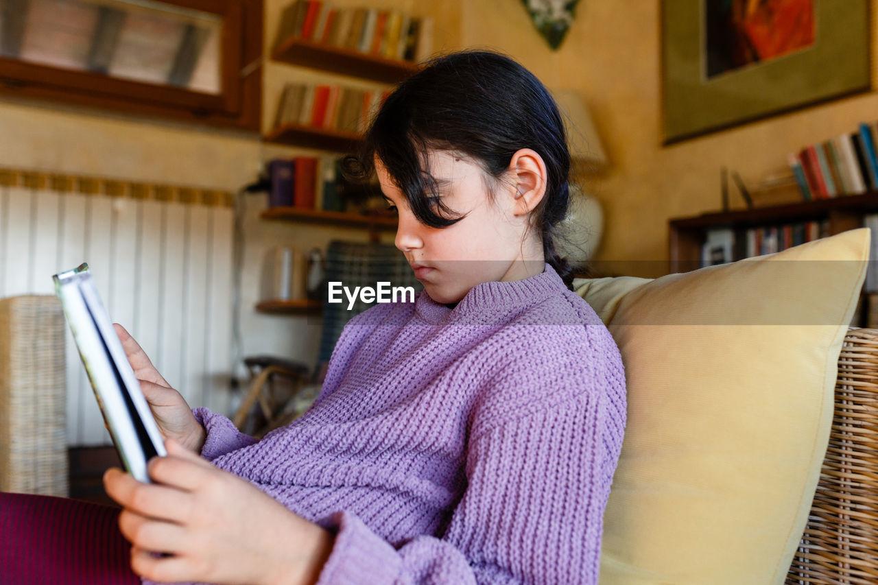 Side view of girl in lilac pullover reading a book in yellow armchair