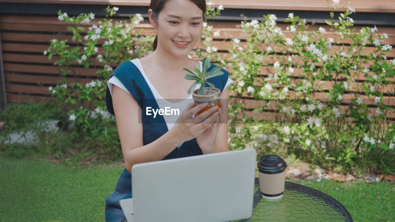 SMILING YOUNG WOMAN USING PHONE WHILE STANDING ON PLANT