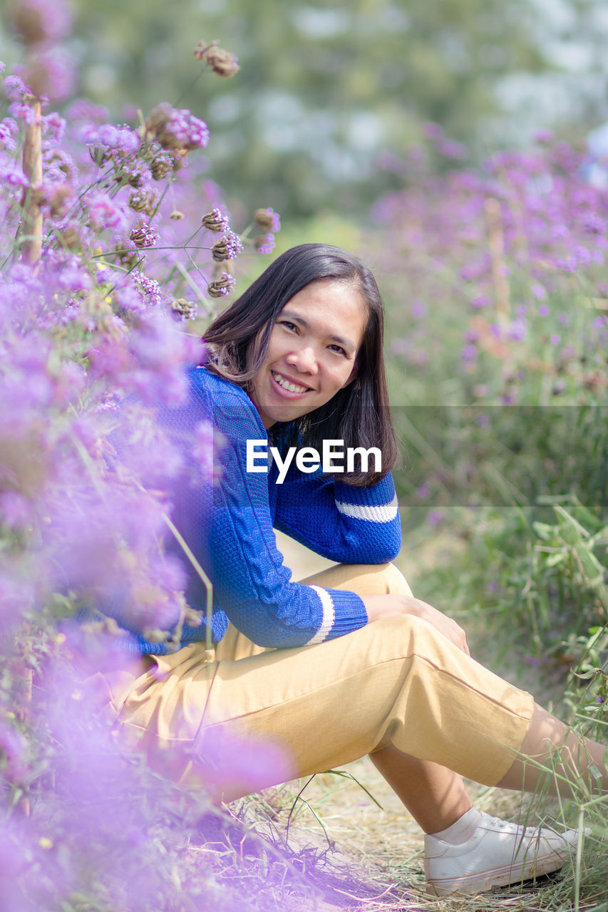 PORTRAIT OF A SMILING YOUNG WOMAN SITTING ON PURPLE FLOWER