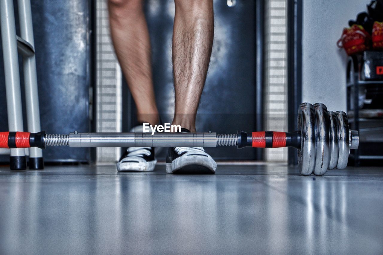 Low section of man standing by dumbbell on tiled floor in gym