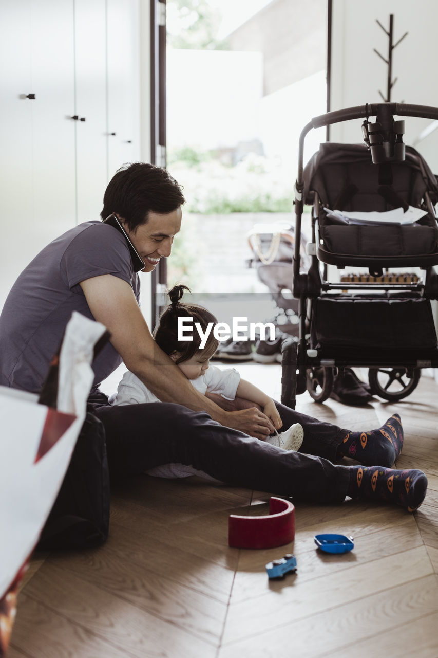 Father talking on smart phone while tying baby son's shoelace at home