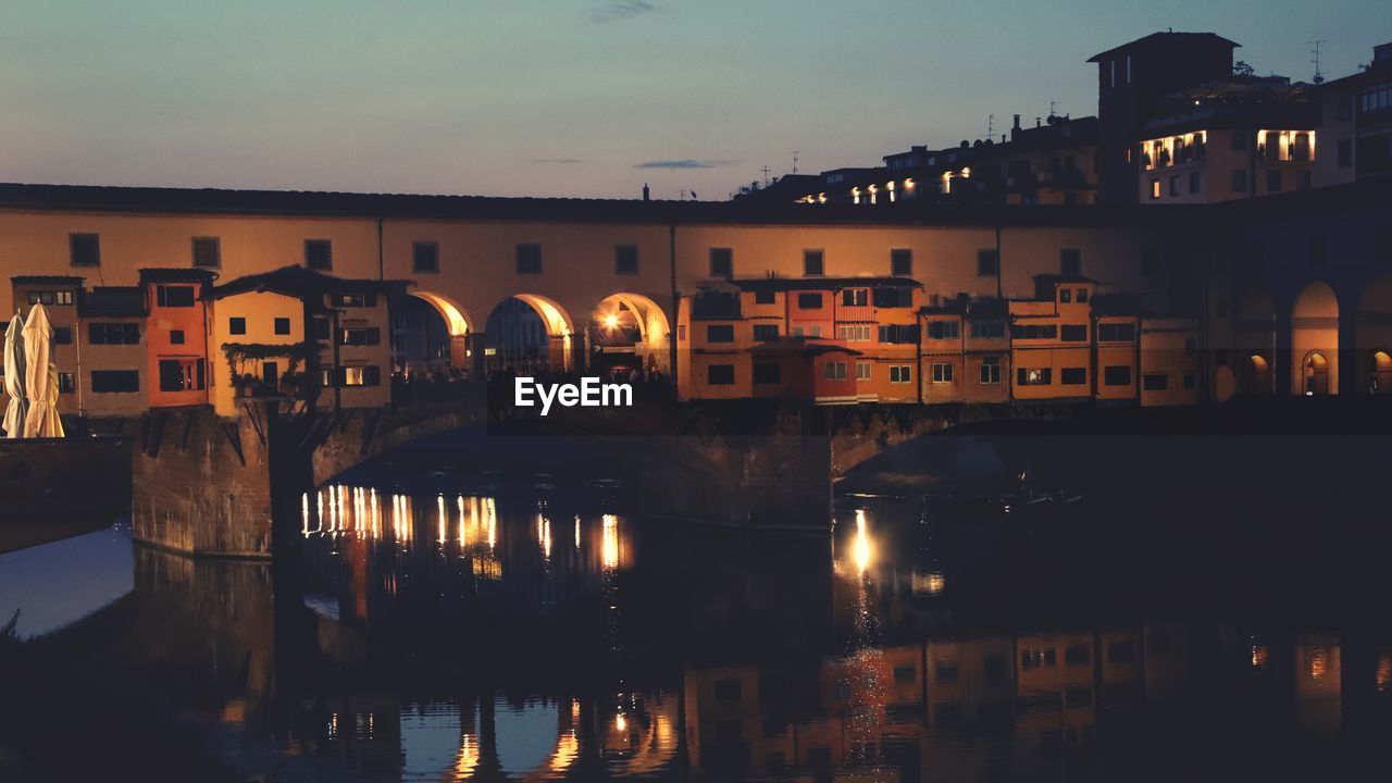 Bridge over river by illuminated buildings in city at dusk