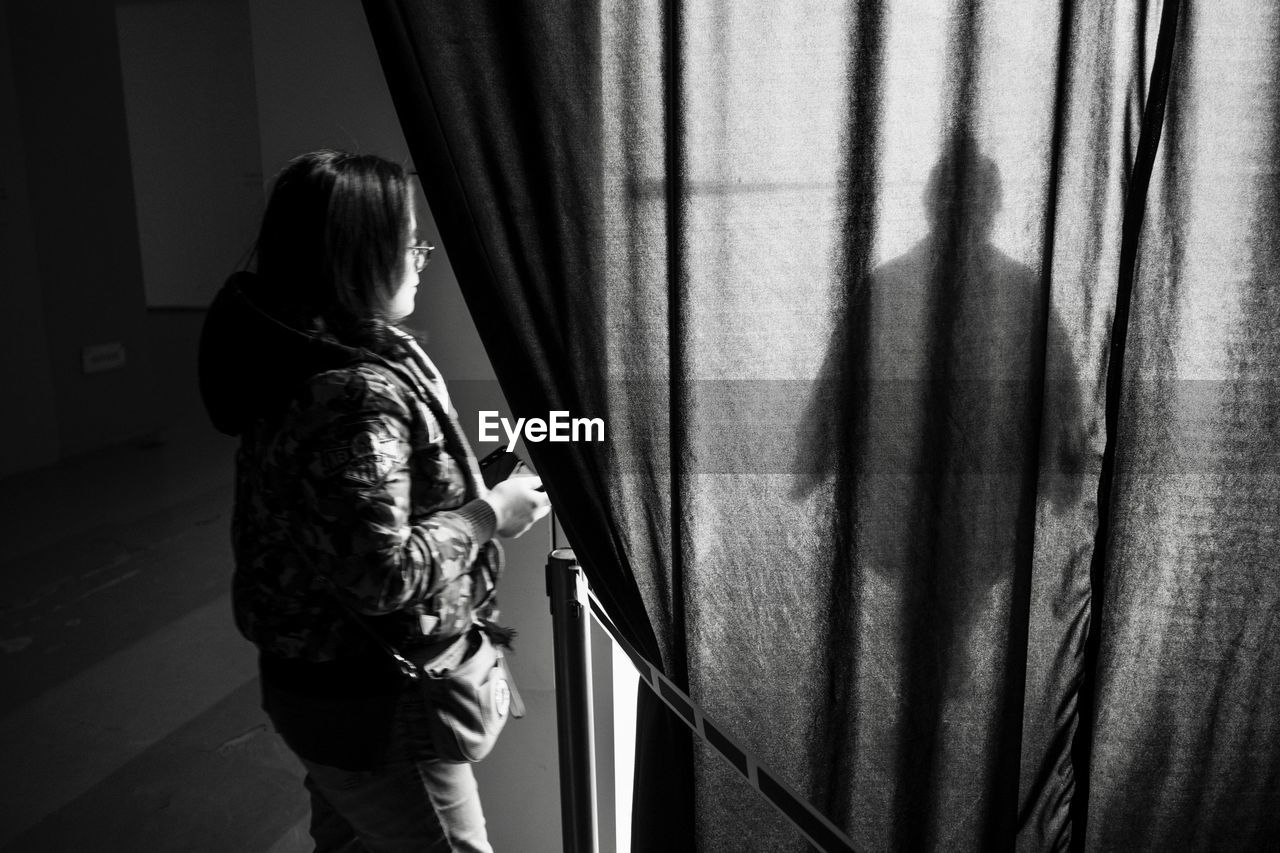 REAR VIEW OF A WOMAN STANDING BY WINDOW