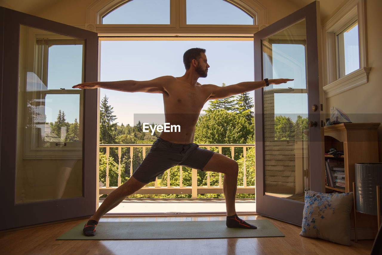 Man doing warrior yoga pose in front of open window doors to forest view.