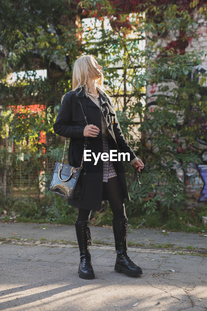 full length, one person, women, adult, blond hair, clothing, footwear, plant, day, spring, nature, standing, lifestyles, coat, walking, casual clothing, leisure activity, hairstyle, tree, outdoors, boot, bag, dress, young adult, long hair, fashion, autumn, handbag, person, fashion accessory, costume, city, female, jacket, rear view, holding, luggage and bags, footpath, looking