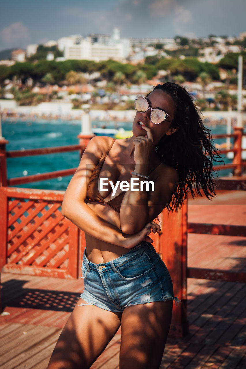 Beautiful young woman wearing sunglasses while standing on pier at beach