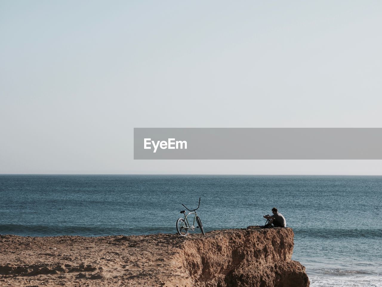 Distant view of man sitting on cliff by sea against clear sky