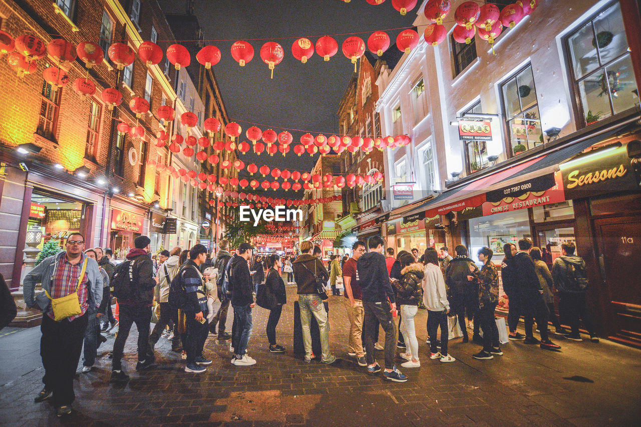 PEOPLE ON ILLUMINATED STREET AMIDST BUILDINGS IN CITY