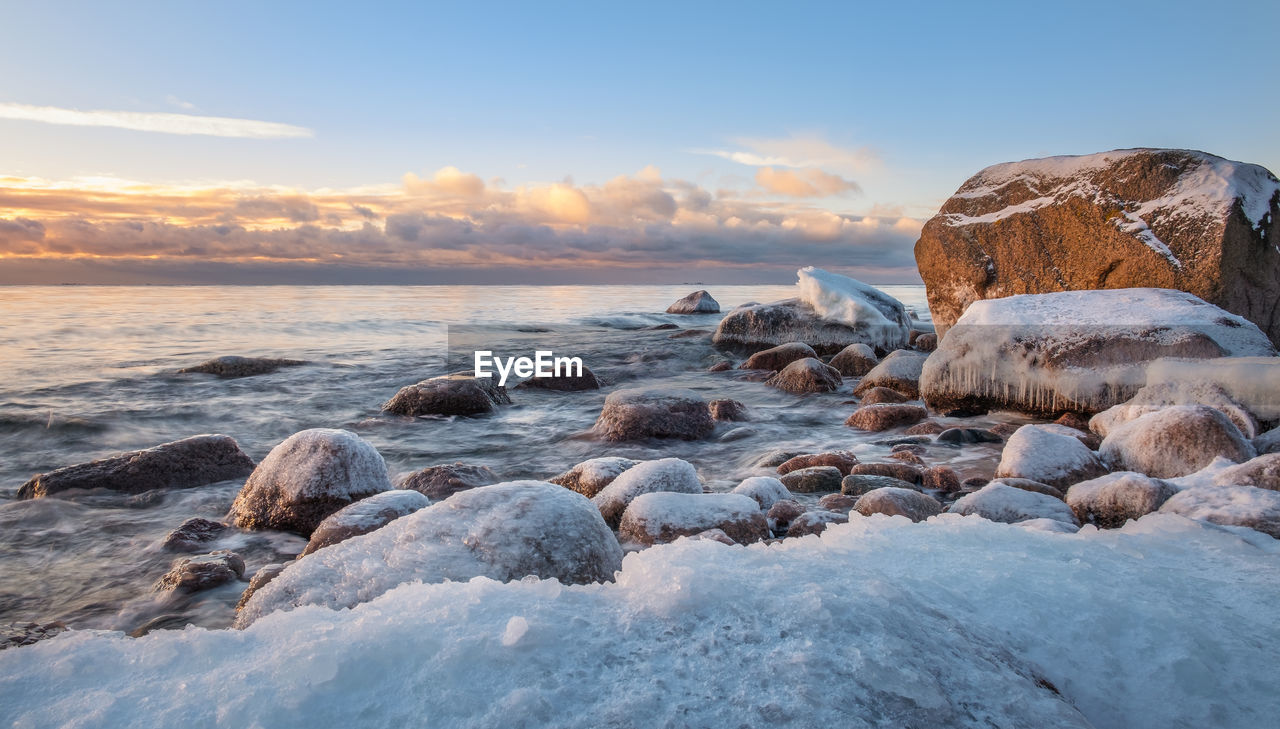 Snow covered rocks at beach against sky during sunset