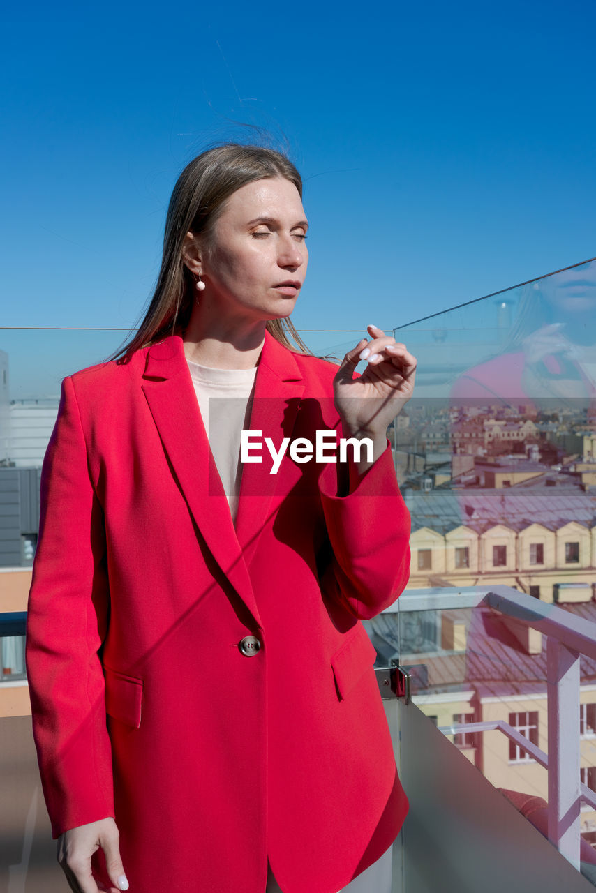 one person, adult, women, architecture, standing, city, sky, businesswoman, young adult, business, clothing, looking, technology, holding, building exterior, waist up, blue, communication, built structure, female, wireless technology, red, day, telephone, coat, nature, person, looking away, cityscape, outdoors, mobile phone, three quarter length, brown hair, smiling, outerwear, travel, clear sky, city life, building, occupation, lifestyles, business finance and industry