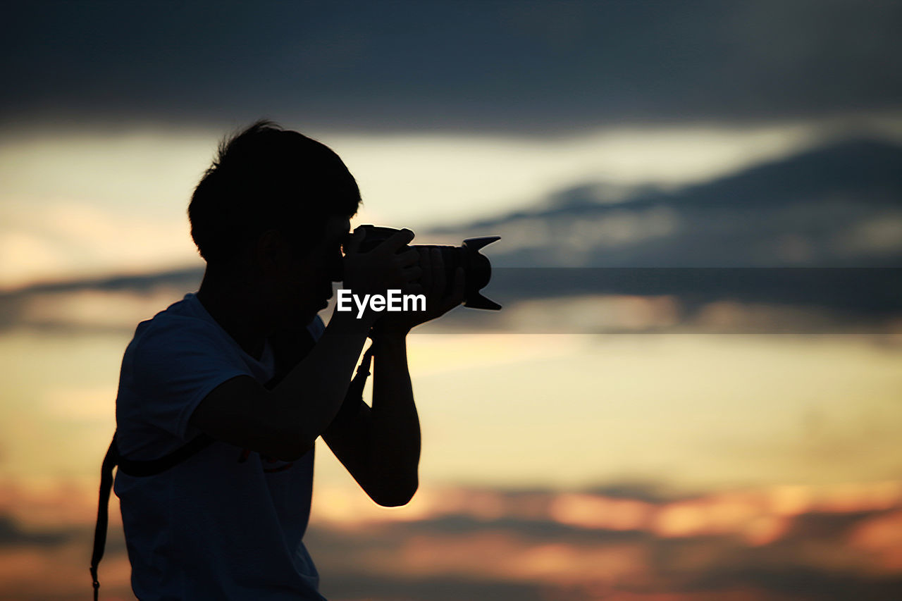 SIDE VIEW OF PERSON PHOTOGRAPHING AGAINST SKY