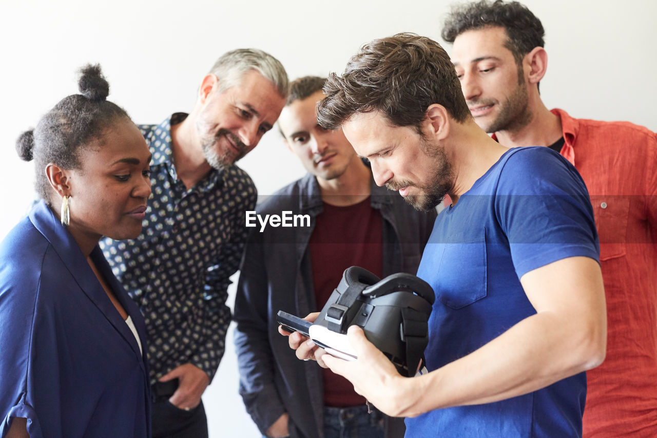 Businessman holding virtual reality simulator while showing mobile phone to colleagues in creative office