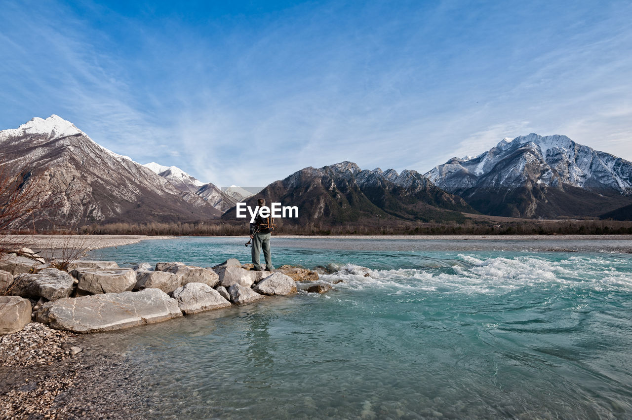 Rear view of man standing on rock by river against sky