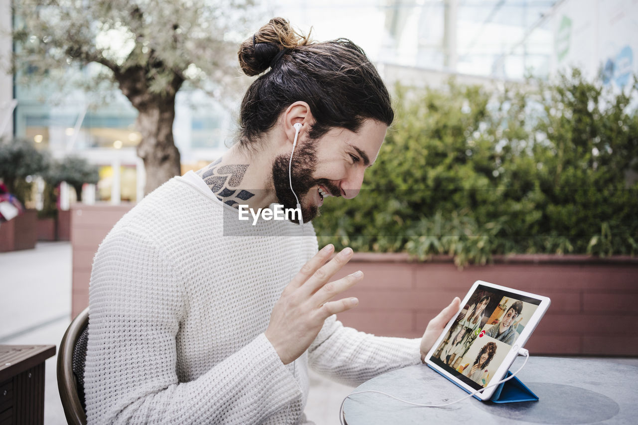 Smiling man waving hand during video call through digital tablet at sidewalk cafe