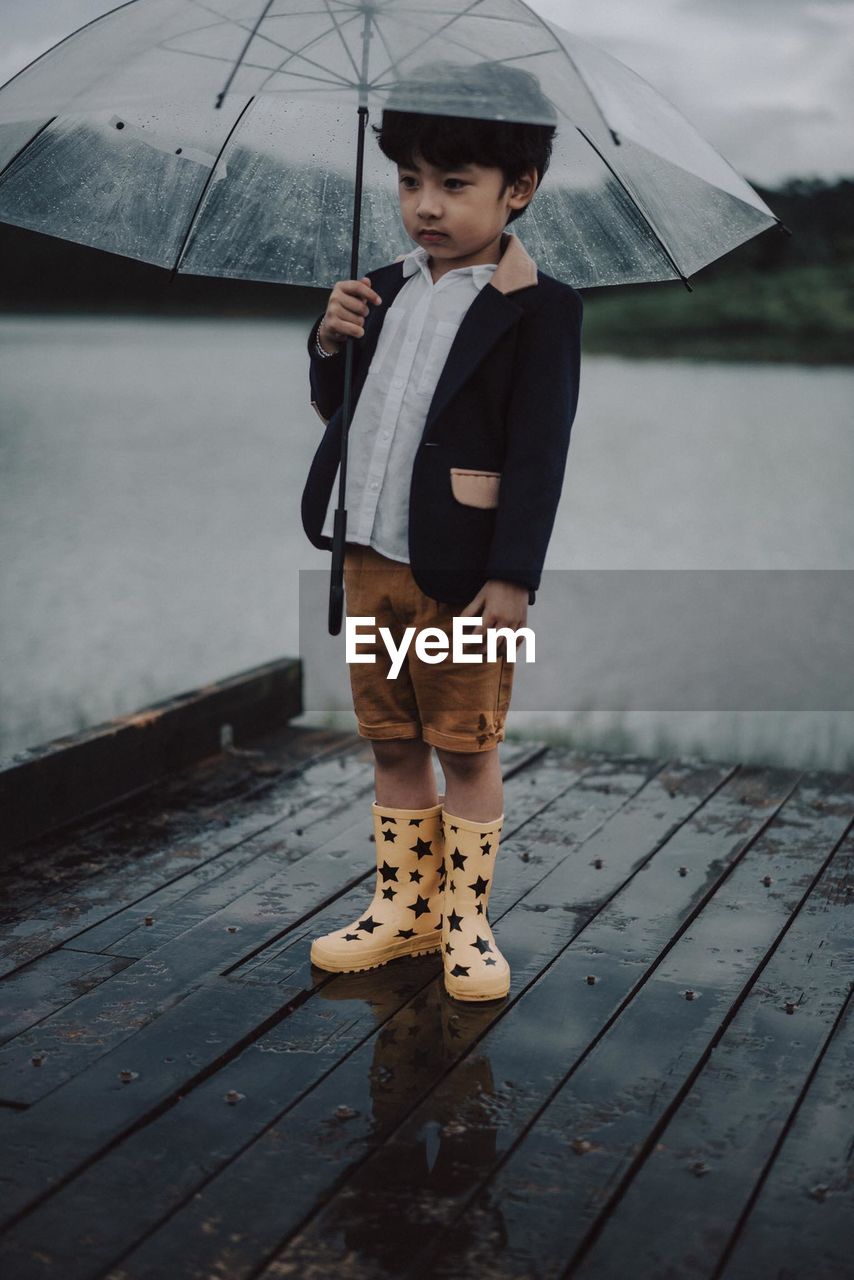 Boy with umbrella standing on pier against lake during rainfall