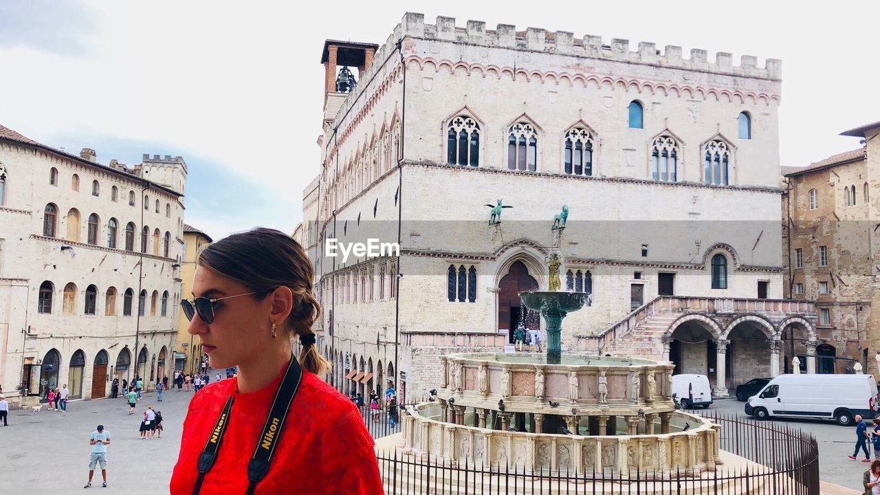 WOMAN STANDING BY BUILDINGS IN CITY AGAINST SKY