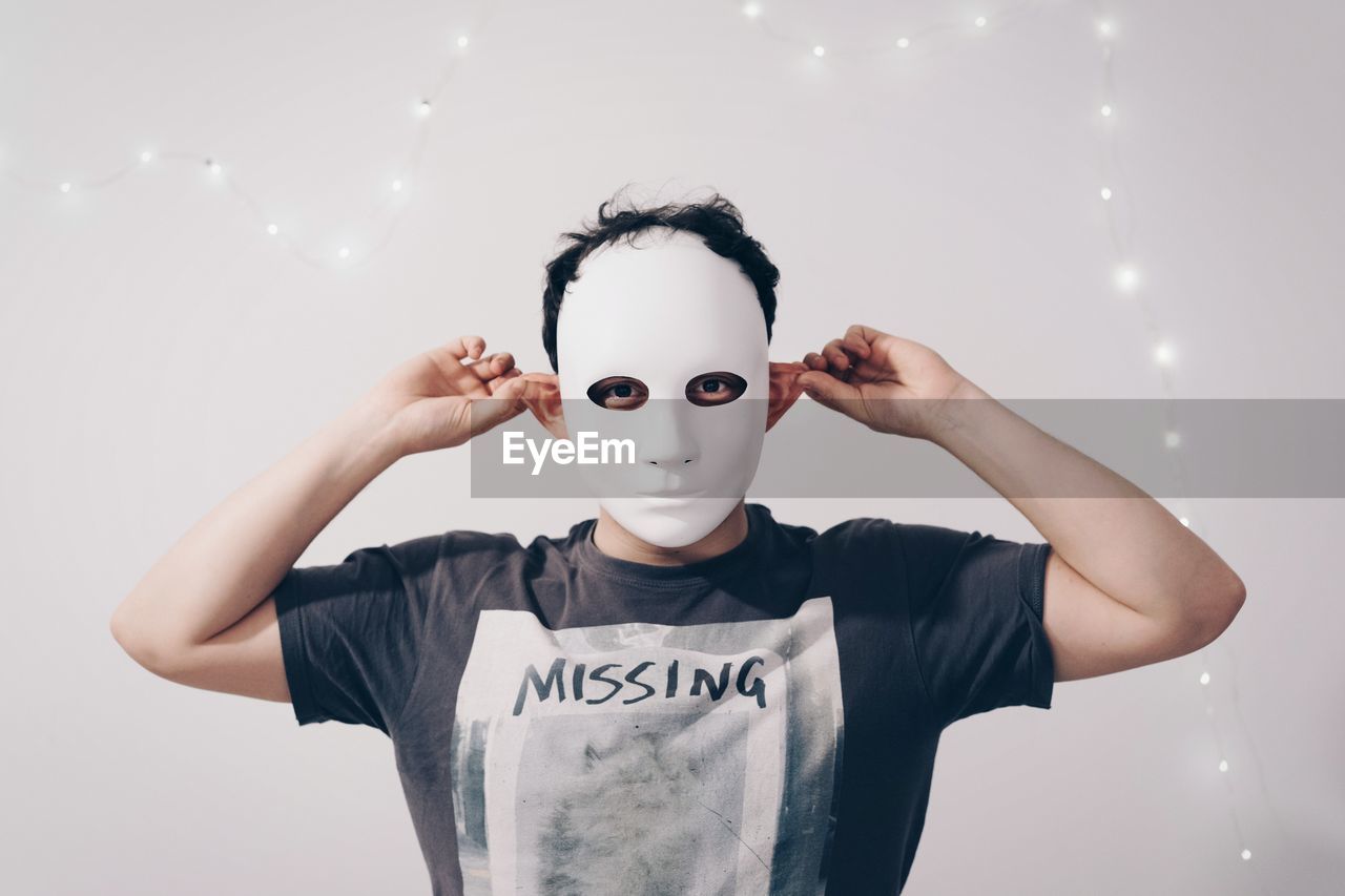 Portrait of man wearing mask while holding ears against string lights decorated white wall