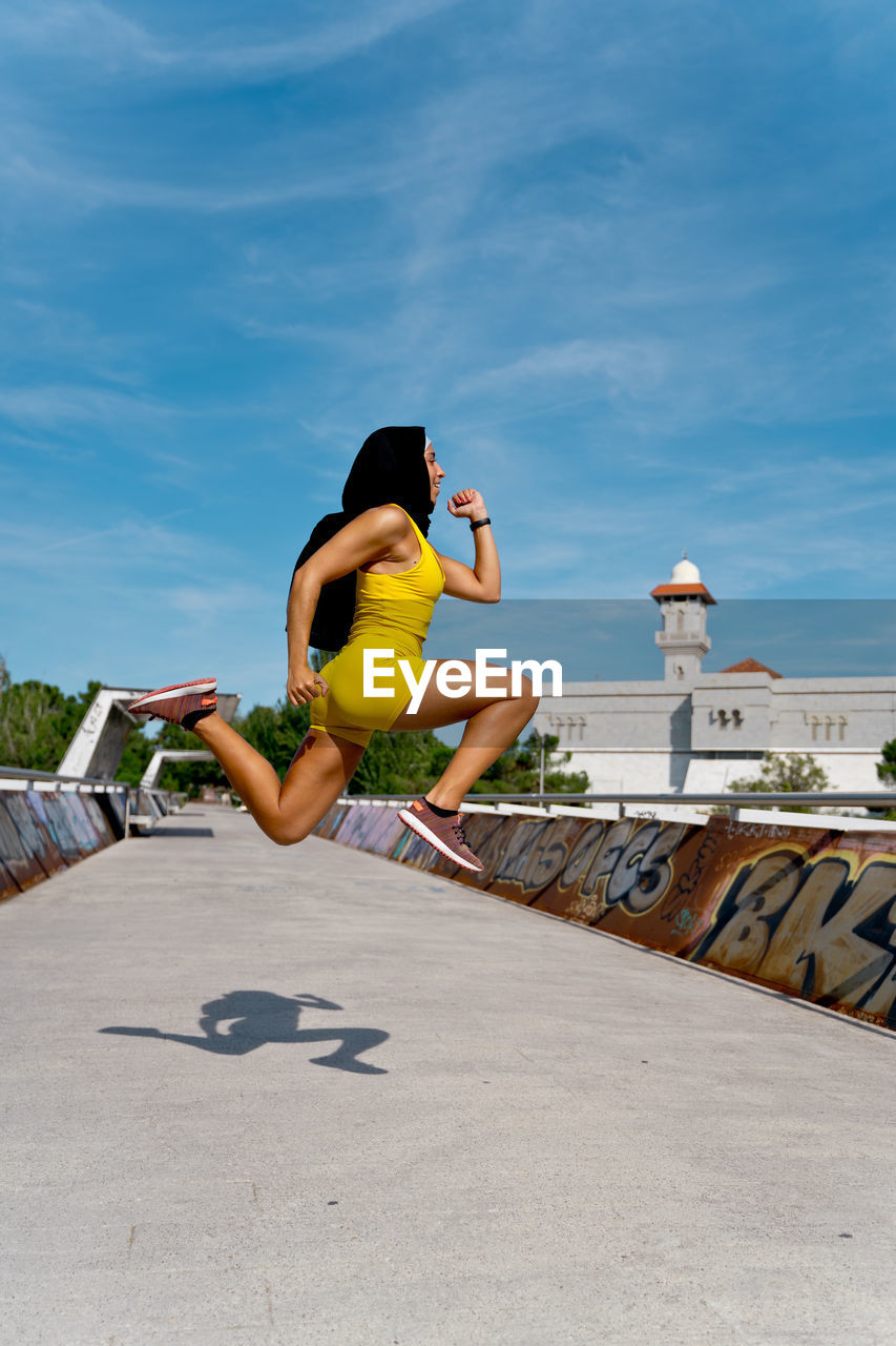 SIDE VIEW OF YOUNG WOMAN JUMPING AGAINST THE SKY