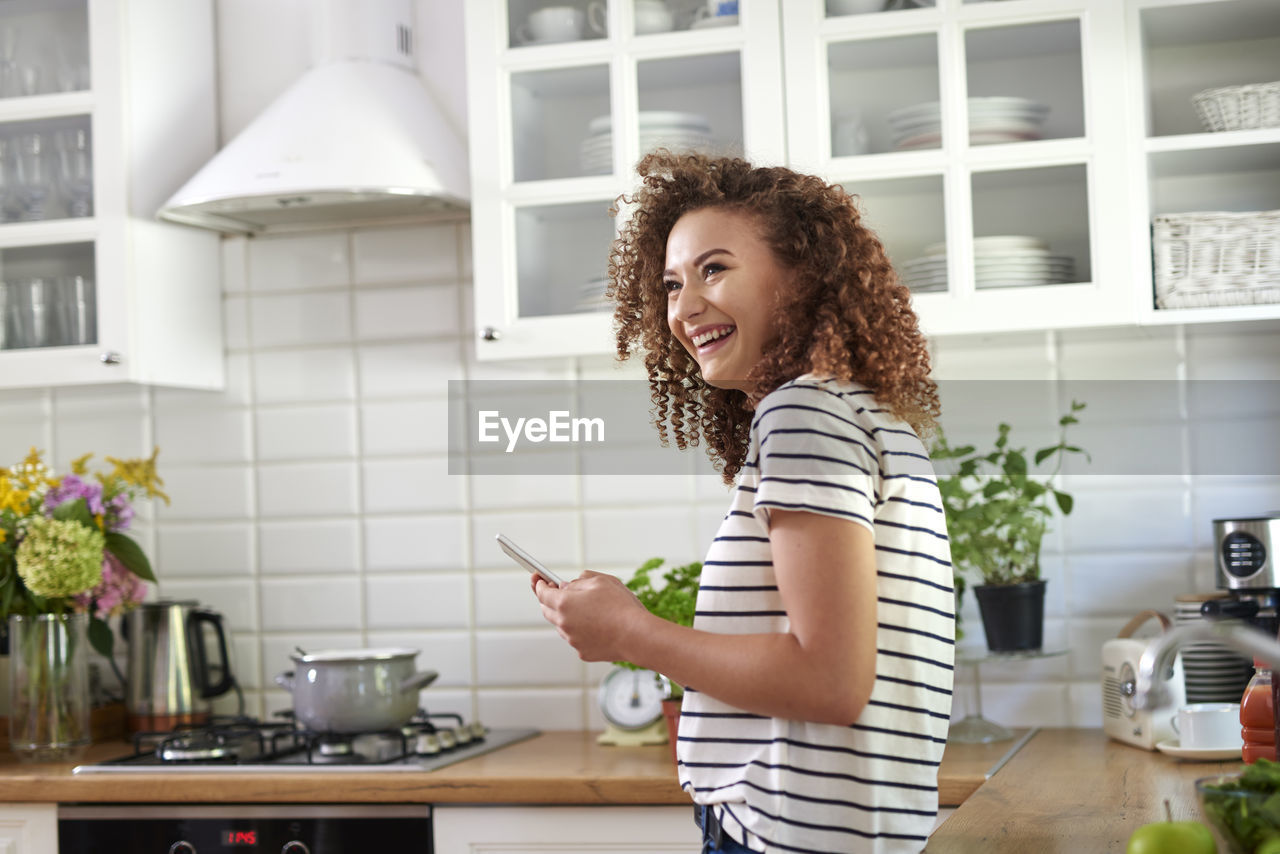 Side view of cheerful teenage girl using smart phone while standing in kitchen at home