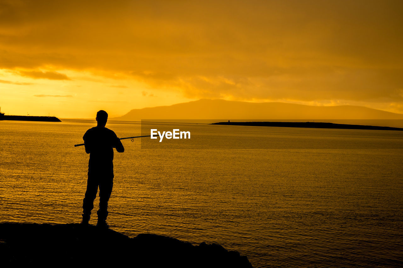 Silhouette man fishing in sea against orange sky