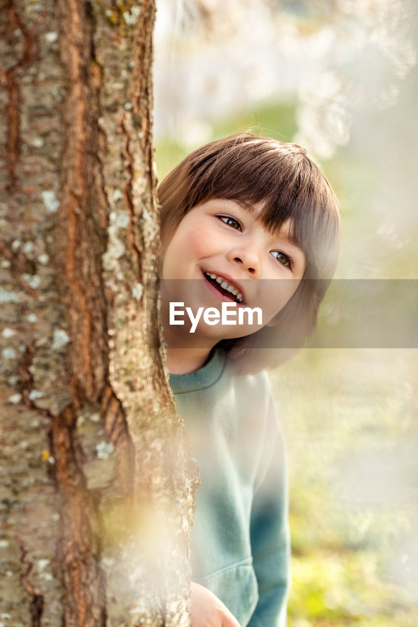 Outdoor portrait of smiling happy toddler boy playing hide and seek hiding behind the tree in park