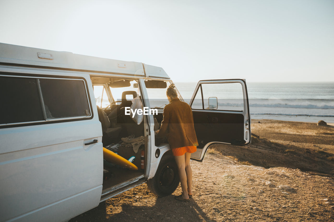 Rear view of woman standing by camping van at beach against clear sky