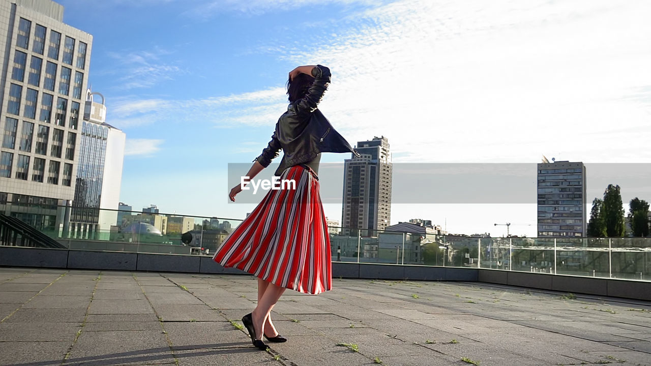 Happy woman wearing striped skirt and jacket on footpath in city