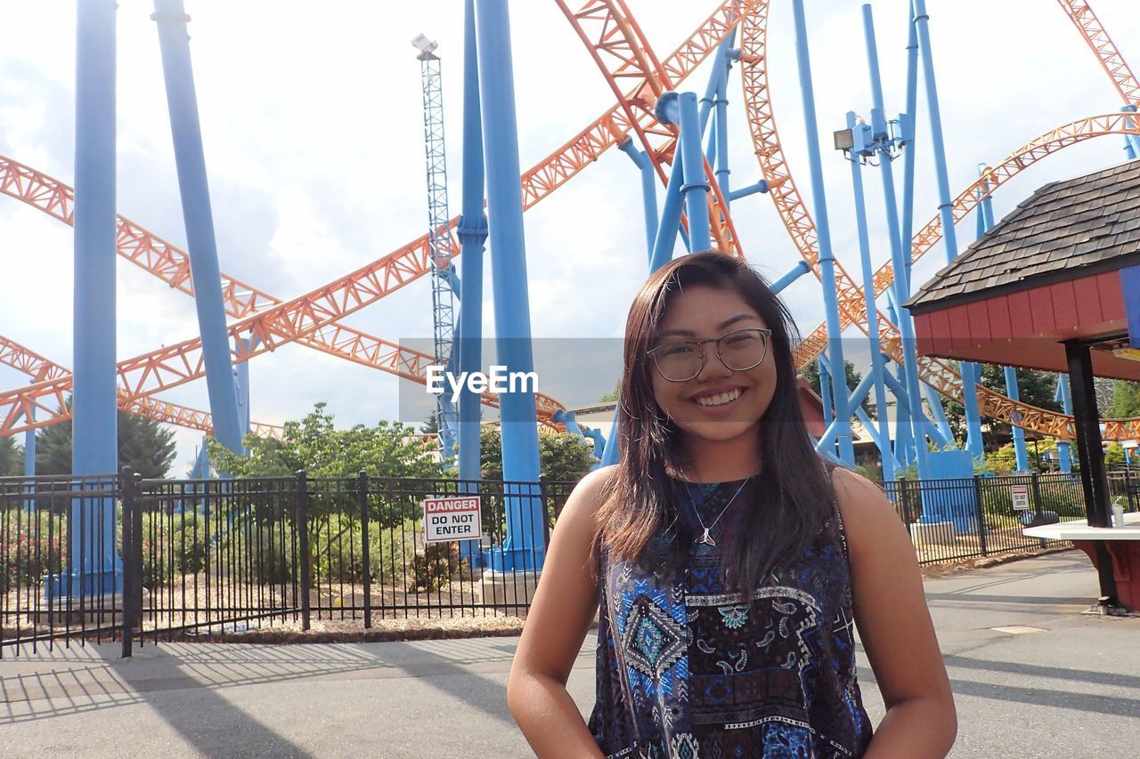 Close-up portrait of woman wearing eyeglasses while standing against rollercoaster