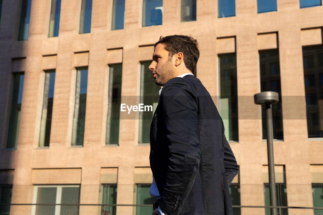 From below side view of thoughtful male in formal clothes looking away with pensive face while standing on sunny street near building with shadow on face