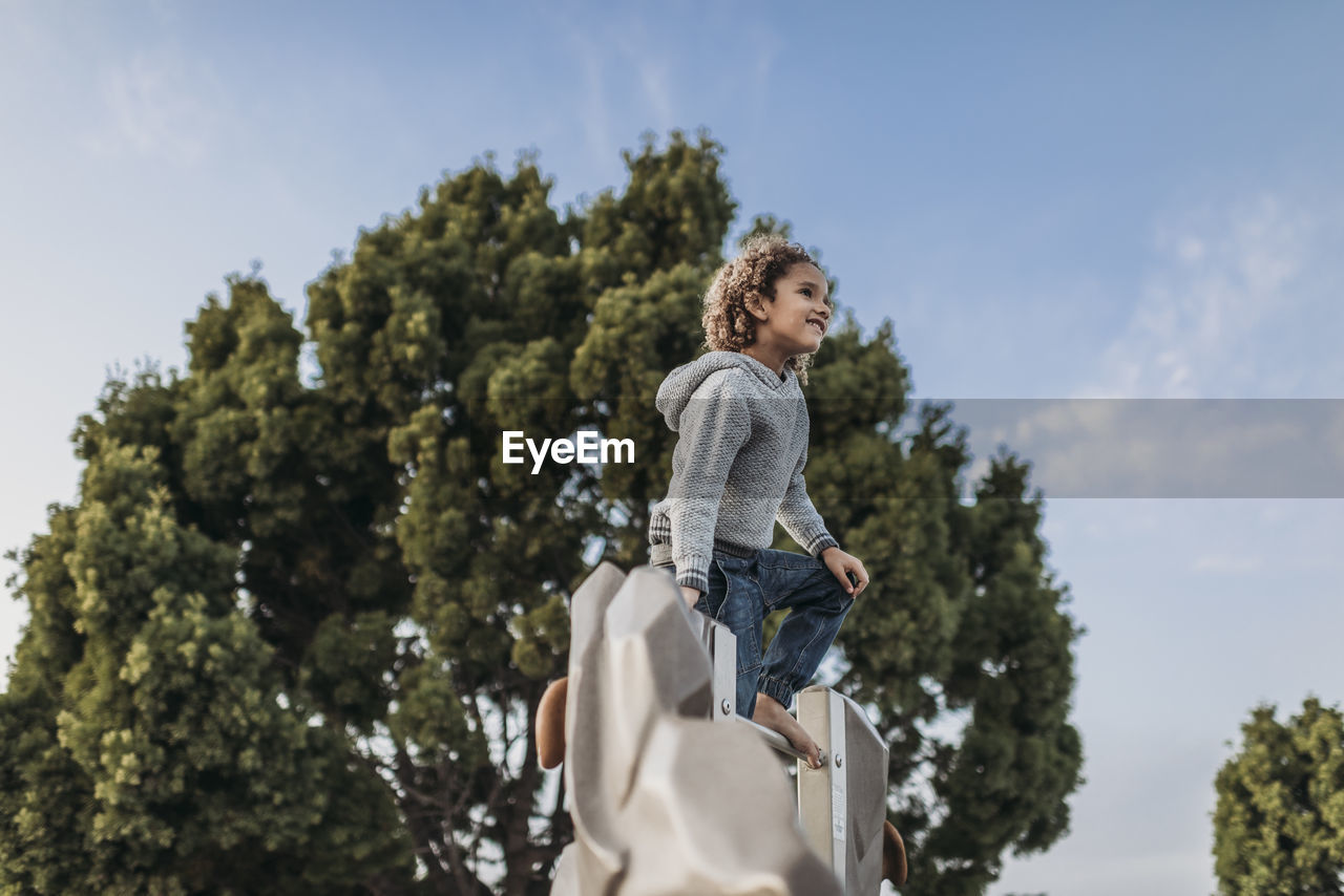 Cute school aged boy standing on top of park structure against trees