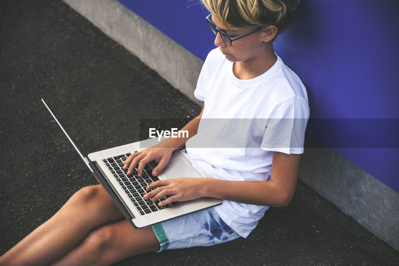 High angle view of boy using laptop while sitting against wall