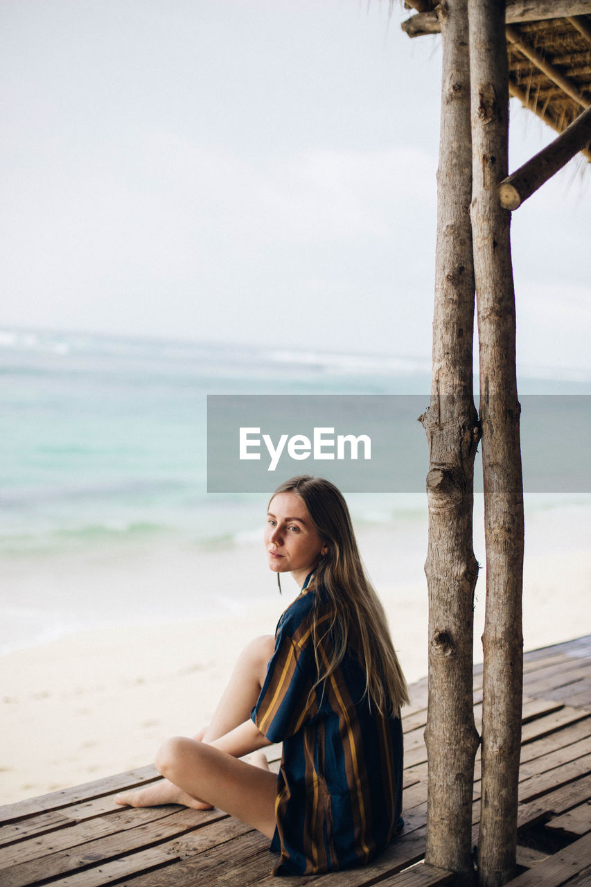 Young woman sitting on shore at beach against sky