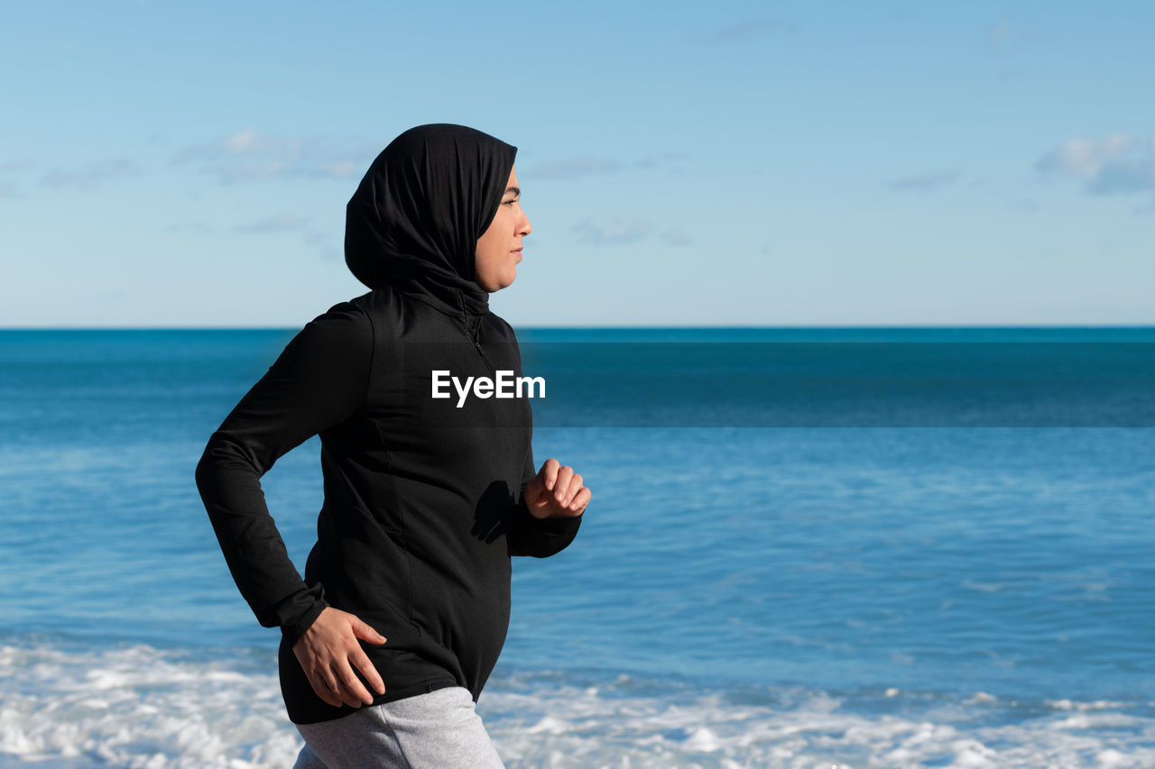 Portrait of young woman standing at beach against sky