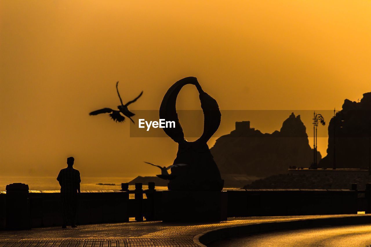 SILHOUETTE MAN STANDING BY SEA AGAINST CLEAR SKY DURING SUNSET