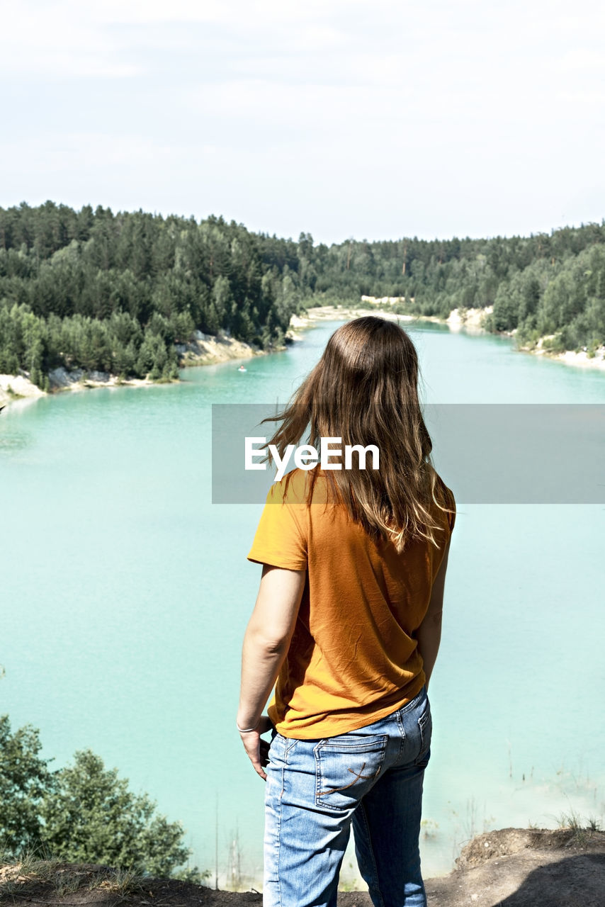 Rear view of blond woman hiker standing on cliff of blue turquoise lake or river and forest, travel