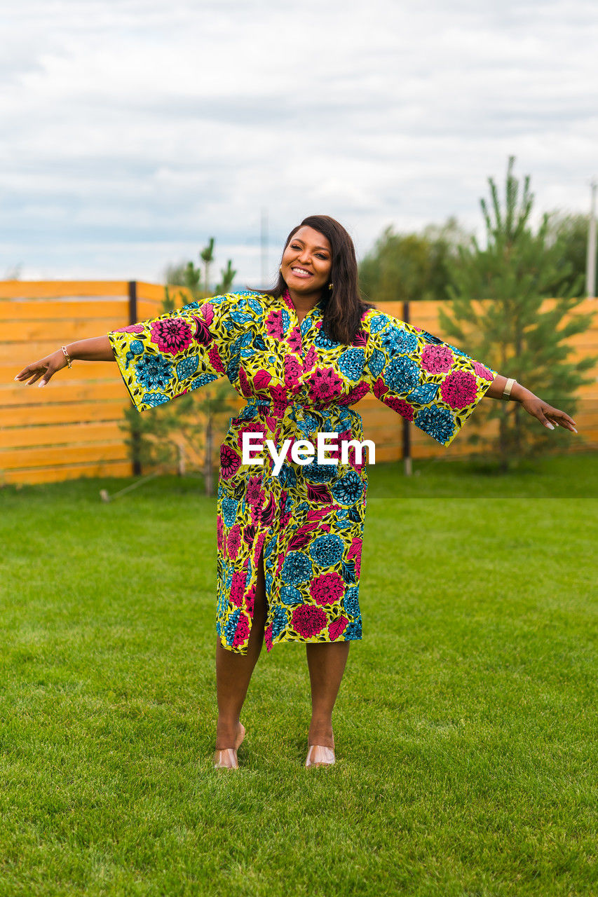 one person, smiling, women, happiness, grass, plant, adult, flower, nature, multi colored, full length, standing, emotion, portrait, young adult, front view, cheerful, clothing, dress, lifestyles, looking at camera, fashion, female, leisure activity, flowering plant, sky, person, outdoors, cloud, day, traditional clothing, holding, field, enjoyment, green, spring, agriculture, fun, land, pattern, hairstyle, smile, yellow, casual clothing, teeth, carefree, arts culture and entertainment, positive emotion, floral pattern