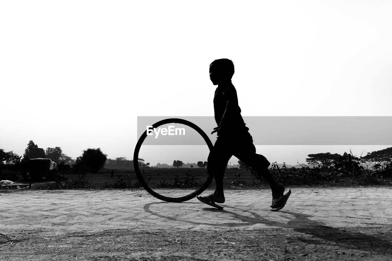 Silhouette boy playing with tire on land against clear sky