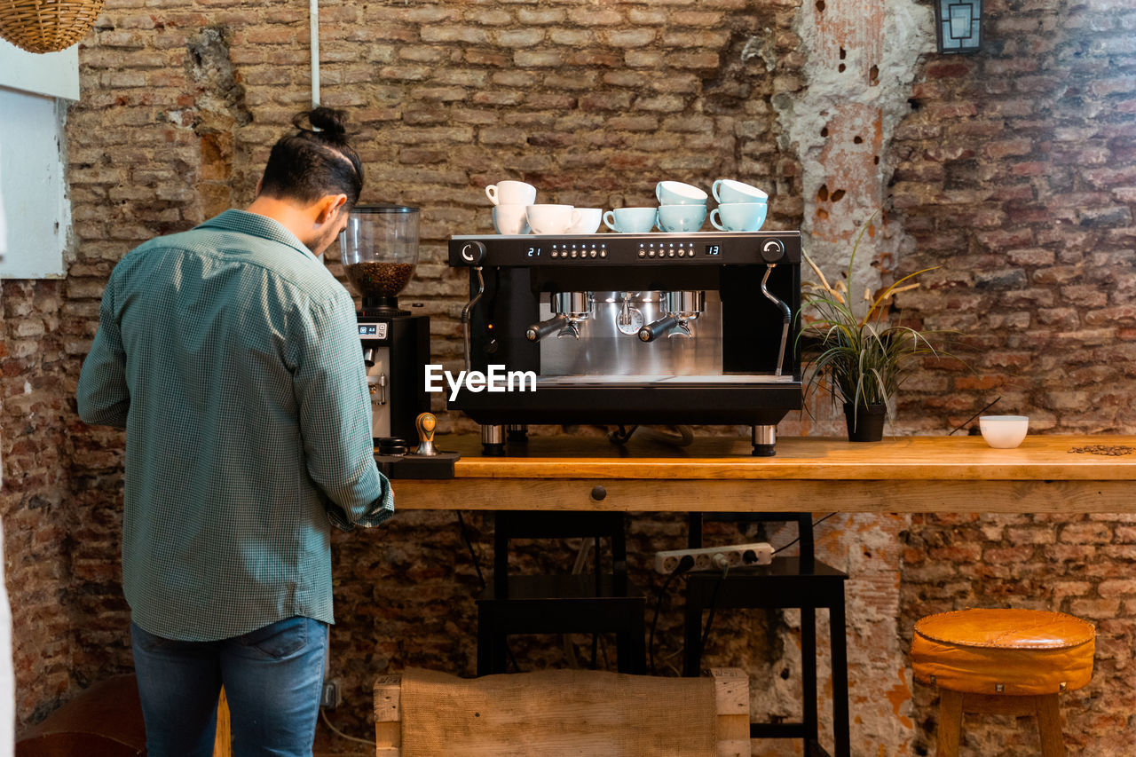 Back view male barista using portafilter and preparing coffee in modern coffeemaker while standing at counter in cafe