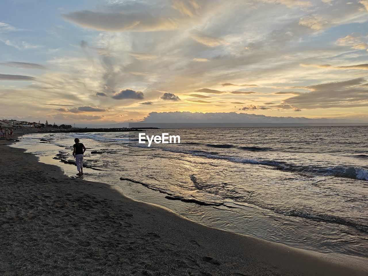 sky, beach, land, sea, water, horizon, shore, ocean, sand, coast, sunset, body of water, beauty in nature, nature, wave, cloud, scenics - nature, full length, one person, sunlight, holiday, leisure activity, tranquility, walking, vacation, trip, environment, dawn, men, adult, travel, landscape, travel destinations, horizon over water, tranquil scene, motion, outdoors, wind wave, lifestyles, coastline, idyllic, sun, reflection, non-urban scene, silhouette