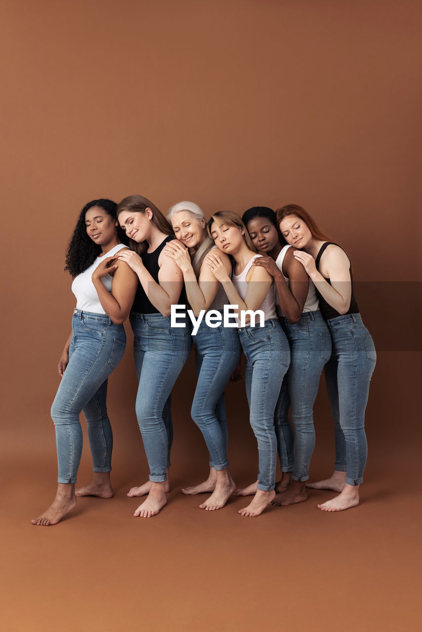 Six multiracial females in casuals hugging together on brown background in studio. 