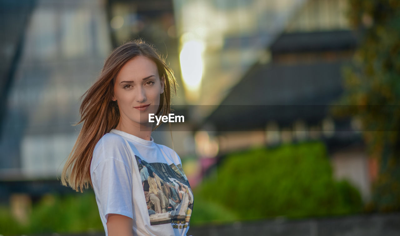 PORTRAIT OF BEAUTIFUL YOUNG WOMAN STANDING AGAINST BLURRED BACKGROUND