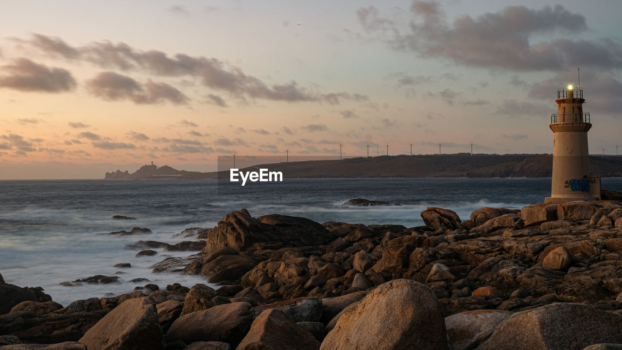 Lighthouse on ocean sea coast rock formation over sunset cloudy sky and wind turbines background