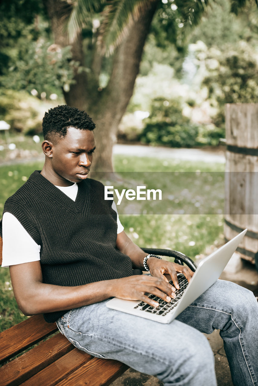 Young black man working on laptop outdoors, looking at screen, focused on work