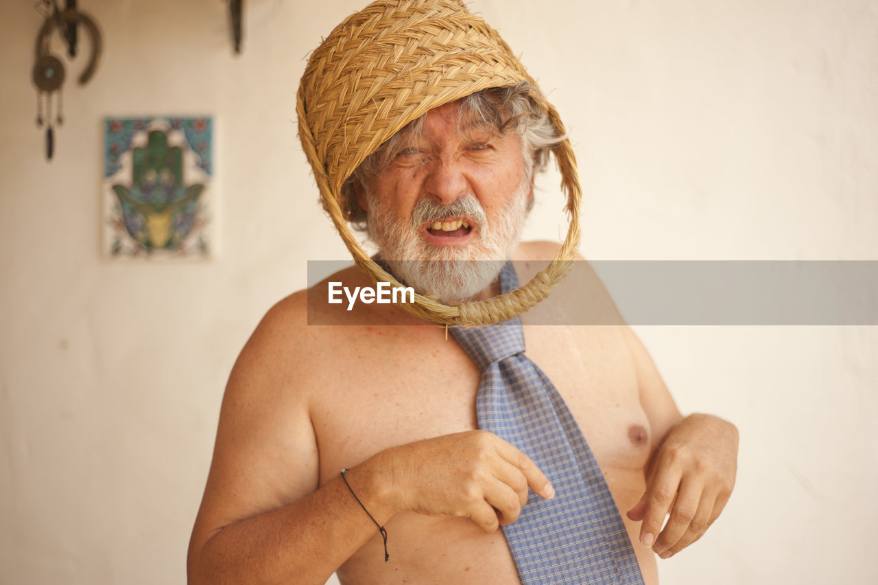 Portrait of shirtless senior man with basket on head standing against wall at home